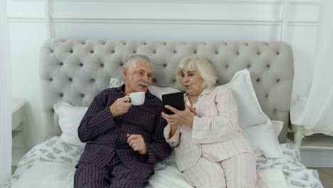 Senior-elderly-couple-wearing-pyjamas-lying-on-bed-looking-on-digital-tablet-laughing-and-having-fun