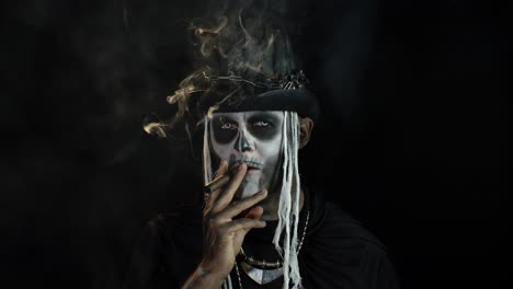 Sinister-man-with-horrible-Halloween-skeleton-makeup-smoking-cigar,-making-faces,-looking-at-camera