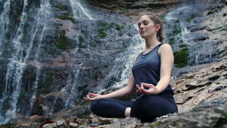 Woman-meditating-by-the-waterfall-and-connecting-with-nature,-low-angle-shot