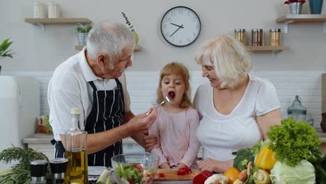 Elderly-grandparents-in-kitchen-feeding-grandchild-girl-with-chopped-red-pepper.-Vegetarian-diet