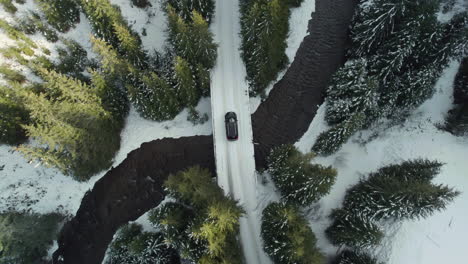 Aerial-view-of-a-black-car-on-a-snowy-road-crossing-over-a-bridge-above-a-mountain-river,-surrounded-by-tall-fir-trees-illuminated-by-sunlight