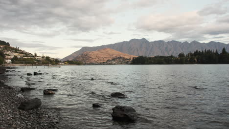 Gentle-waves-against-rocky-Queenstown-beach-as-dusk-settles,-framing-picturesque-view-of-distant-mountains