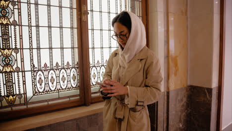 Young-girl-scrolling-on-her-phone-and-standing-next-to-a-window-wearing-a-hoodie-and-a-beige-trench-coat