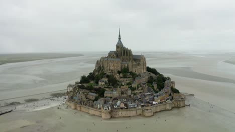 mont-saint-michel-drone-camera-going-towards-the-castle,-low-rise-there-are-many-trees-around-the-buildings