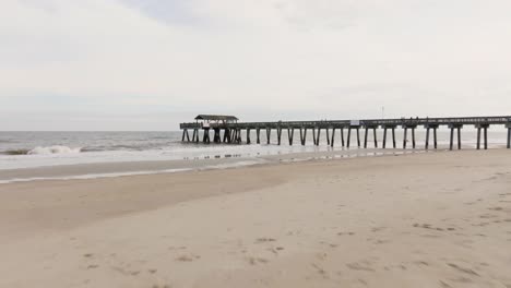 Drone-of-an-empty-beach-in-Tybee-Island-approaching-pier-and-ocean-with-rough-waves,-low-to-the-sand