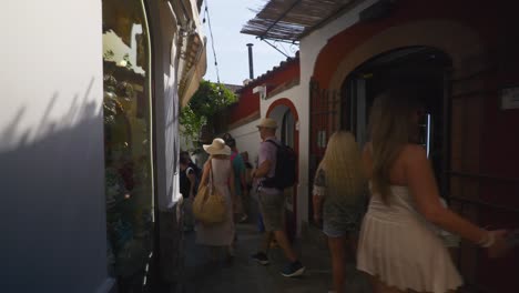Amalfi-Positano-Italy-Immersive-Travel-Tourism-Mediterranean-Sea-Coast-Water-Europe,-Walking,-4K-|-Moving-Through-A-Tightly-Crowded-Dark-Walk-Alley-with-Green-Vines-Overhead-Behind-Blonde-Haired-Woman