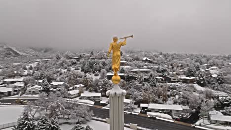 Drone-circles-around-golden-angel-with-trumpet-with-snow-covered-neighborhood-behind