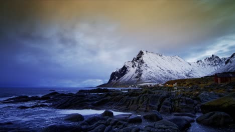 Floating-clouds-reflecting-sunset-moving-from-the-background-towards-the-foreground-above-snow-covered-mountain-behind-the-village-by-the-rocky-shore