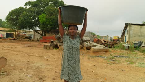 Walking-while-carrying-a-water-filled-basin-on-top-of-her,-a-local-woman-is-on-her-way-back-to-her-house-in-a-neighborhood-in-Kumasi,-Ghana,-in-Africa