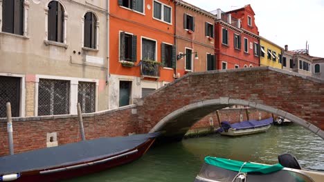Frau-überquert-Eine-Venezianische-Backsteinbrücke-Mit-Im-Kanal-Geparkten-Lokalen-Motorbooten