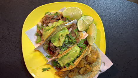 Slow-motion-overhead-shot-of-a-yellow-plastic-plate-with-a-assorted-traditional-mexican-tacos-served-with-slices-of-lime-and-guacamole-on-top