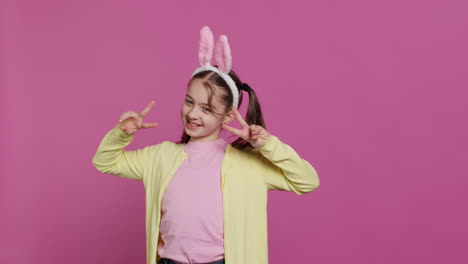 Young-cheerful-kid-with-pigtails-showing-peace-sign-in-studio