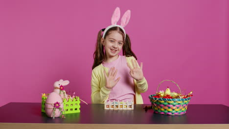 Joyful-child-putting-bunny-ears-on-her-head-and-waving-at-camera