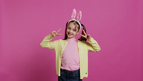 Young-cheerful-kid-with-pigtails-showing-peace-sign-in-studio