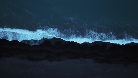Cold-ocean-waves-lapping-a-black-volcanic-sand-beach,-aerial-directly-above-shot