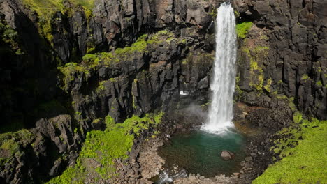 Waterfall-with-clear-water-pouring-down-black-volcanic-stone-cliff,-aerial-view