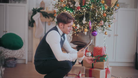 Mann-Nutzt-Digitales-Tablet-Für-Videoanrufe,-Während-Er-Zu-Hause-Weihnachtsgeschenke-Arrangiert