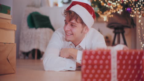Cheerful-man-in-Santa-hat-sliding-Christmas-presents-while-lying-on-floor