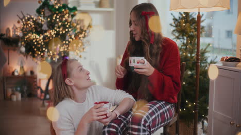 Fröhliche-Schwestern-Reden,-Während-Sie-Zu-Weihnachten-Kaffeetassen-Auf-Einem-Stuhl-Halten