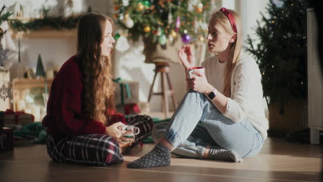 Schöne-Schwestern-Mit-Kaffeetassen,-Die-Zu-Weihnachten-Auf-Dem-Boden-Zu-Hause-Reden