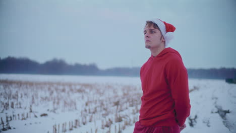 Thoughtful-man-in-red-hooded-jacket-and-Santa-hat-standing-on-snow-covered-landscape