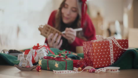 Weihnachtsgeschenke-Und-Ornamente-Auf-Dem-Boden-Mit-Einer-Frau,-Die-Zu-Hause-Ein-Papphaus-Ausmalt