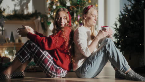Happy-sisters-enjoying-coffee-while-sitting-on-floor-at-home-during-Christmas