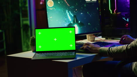 Green-screen-laptop-next-to-man-having-fun-by-using-gaming-keyboard-to-fly-spaceship-in-SF-videogame