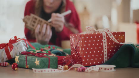 Verpackte-Weihnachtsgeschenke-Auf-Dem-Boden-Mit-Frau,-Die-Zu-Hause-Ein-Papphaus-Ausmalt
