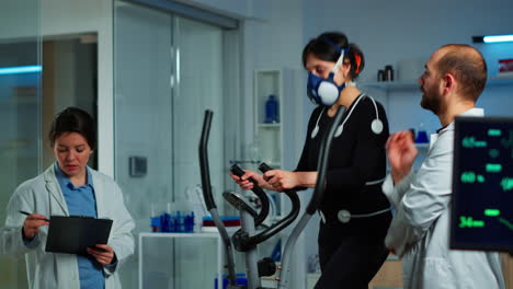 Doctor-witing-on-clipboard-while-patient-with-mask-running-on-cross-trainer
