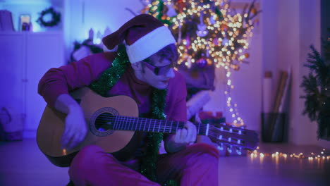 Aufgeregter-Mann-Spielt-Gitarre,-Während-Er-Zu-Weihnachten-Auf-Dem-Boden-Eines-Beleuchteten-Hauses-Sitzt