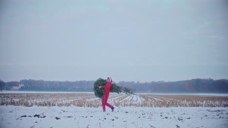 Cheerful-man-carrying-Christmas-tree-on-snow-covered-landscape