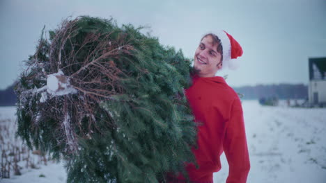 Happy-man-carrying-Christmas-tree-while-walking-on-snow-covered-field