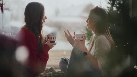 Schwestern-Reden,-Während-Sie-Zu-Weihnachten-Zu-Hause-Kaffeetassen-Am-Fenster-Halten