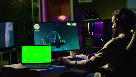 Green-screen-laptop-next-to-man-using-gaming-keyboard-to-play-spaceship-flying-singleplayer-game