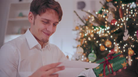 Lächelnder-Mann-Hält-Ein-Weihnachtsgeschenk-In-Der-Hand,-Während-Er-Zu-Hause-Einen-Brief-Liest