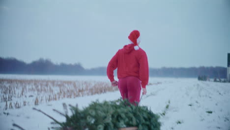 Happy-man-pulling-Christmas-tree-while-running-on-snow-covered-landscape