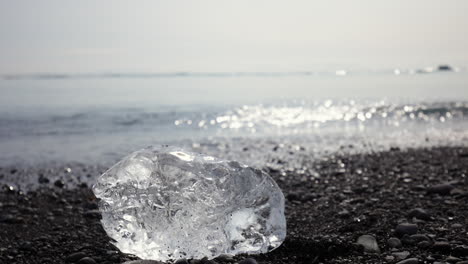 Iceberg-chunk-on-a-black-sand-beach,-sparkling-ocean-in-the-background,-close-up