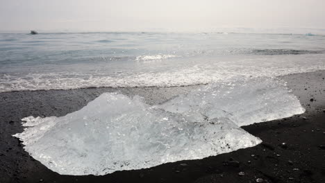 Ocean-waves-lapping-up-chunks-of-ice-sparkling-on-a-black-sand-beach,-handheld