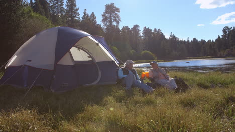 Senior-couple-sitting-outside-tent-drinking-near-a-lake