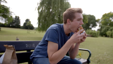 Young-white-man-sitting-on-a-park-bench-enjoying-a-burger