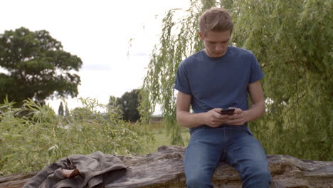 Junger-Mann-Sitzt-Auf-Einem-Umgestürzten-Baum-Und-Sendet-Nachrichten-Mit-Dem-Smartphone