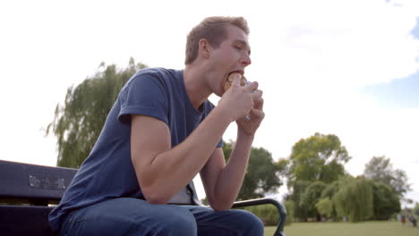 Young-man-sitting-on-a-park-bench-eating-burger,-low-angle