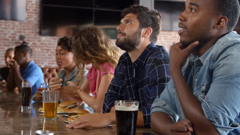 Group-Of-Friends-Watching-Game-In-Sports-Bar-On-Screens
