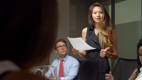 Asian-woman-stands-to-address-colleagues-at-business-meeting