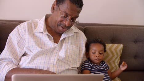 Grandfather-And-Grandson-Sit-On-Sofa-At-Home-Using-Laptop