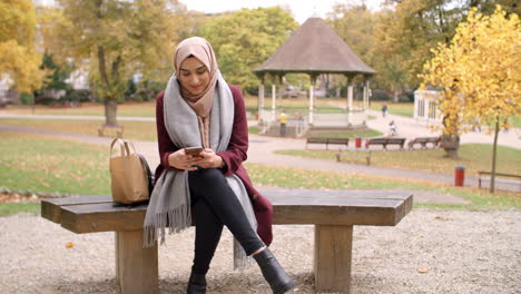 Mujer-Musulmana-Británica-Enviando-Mensajes-De-Texto-Por-Teléfono-Móvil-En-El-Parque