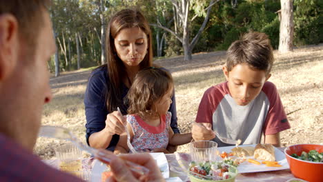 Mixed-race-family-eat-together-at-a-picnic-table-in-a-park