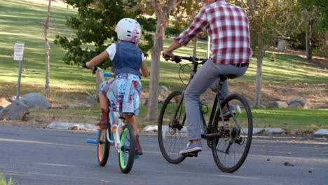 Father-and-daughter-riding-bikes-together-in-a-park,-pan
