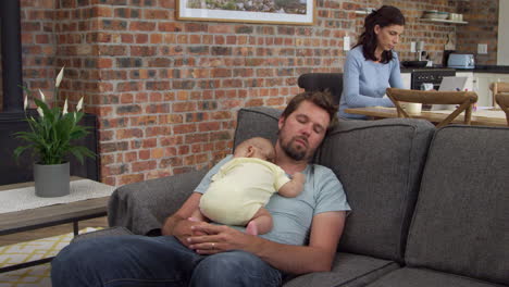 Father-With-Baby-Son-Sleep-On-Sofa-As-Mother-Works-On-Laptop